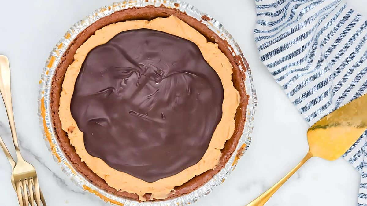 A chocolate pie with a smooth, dark chocolate topping in a foil-lined crust on a marble surface. A blue-striped cloth is beside it, with a gold pie server and two forks nearby.