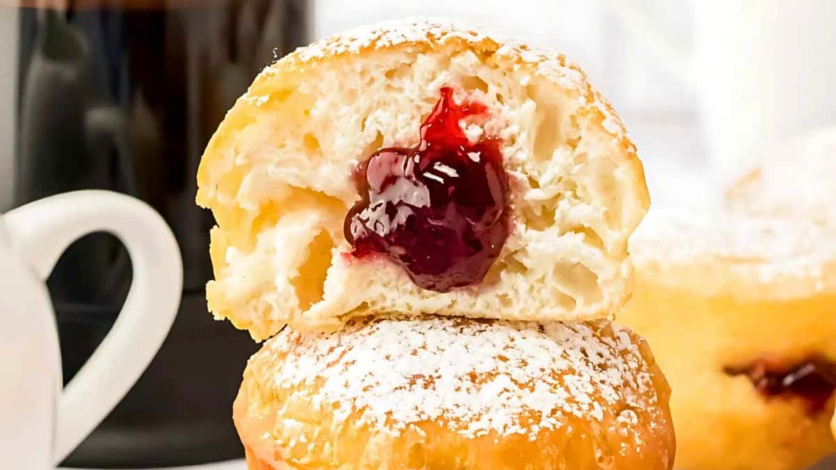 A close-up of a pastry dusted with powdered sugar, filled with glossy red jam, is stacked on another pastry. A cup of coffee is partially visible in the background.