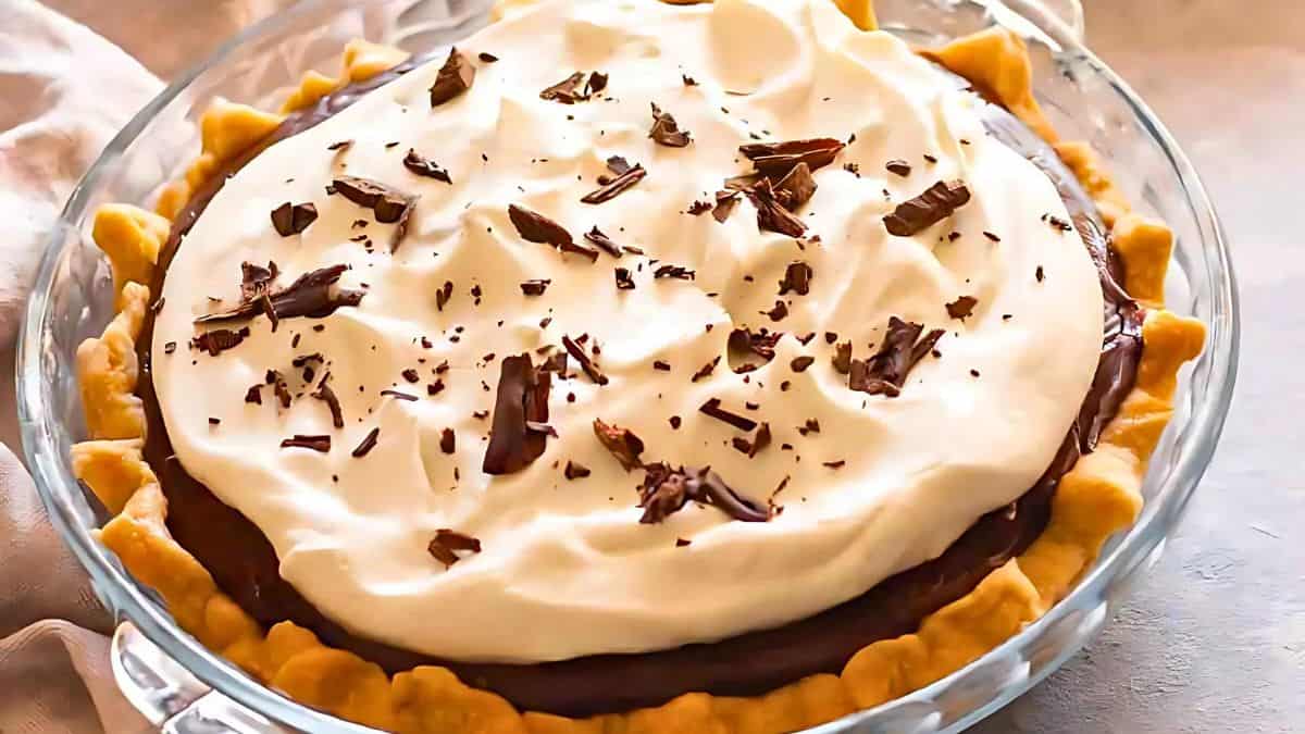 A chocolate cream pie in a glass dish, topped with whipped cream and chocolate shavings. The pie has a golden, crimped crust and is set on a light-colored surface.