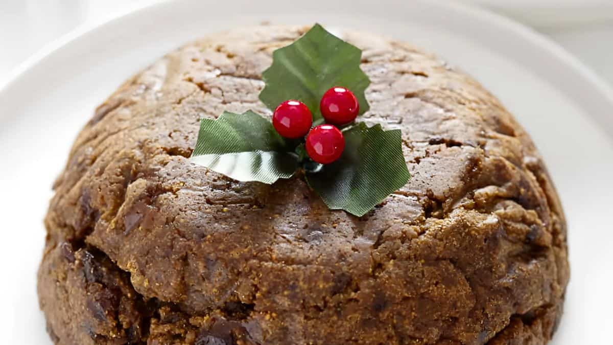A traditional Christmas pudding on a white plate, topped with decorative green holly leaves and three red berries.