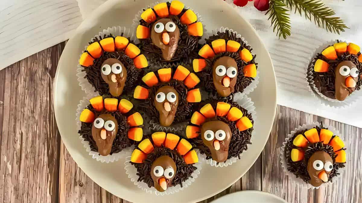 A plate of turkey-themed cupcakes decorated with chocolate frosting, candy eyes, and candy corn arranged as feathers, resembling turkeys. The cupcakes are placed in paper liners on a wooden table.