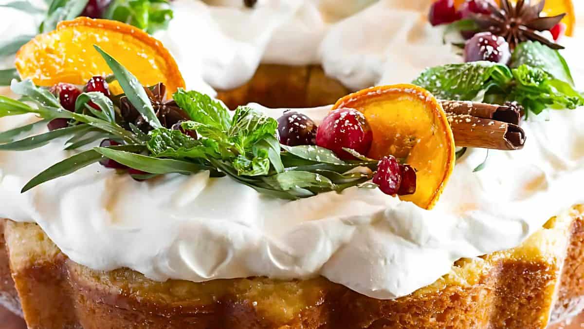 A close-up of a frosted cake decorated with whipped cream, orange slices, cinnamon sticks, mint leaves, cranberries, and star anise. The festive toppings give it a holiday feel.