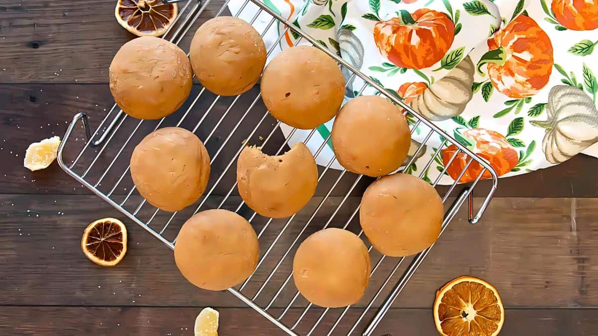 Nine round cookies or pastries on a cooling rack. One has a bite taken out. They are on a wooden table, surrounded by dried orange slices. A white cloth with orange fruit patterns is beside them.