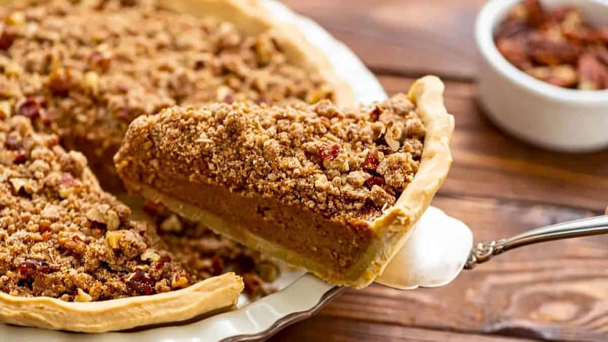 A slice of pie is being lifted from a whole pie. The pie has a golden, flaky crust and is topped with a crumbly pecan streusel. In the background, there is a small bowl filled with pecans on a wooden table.