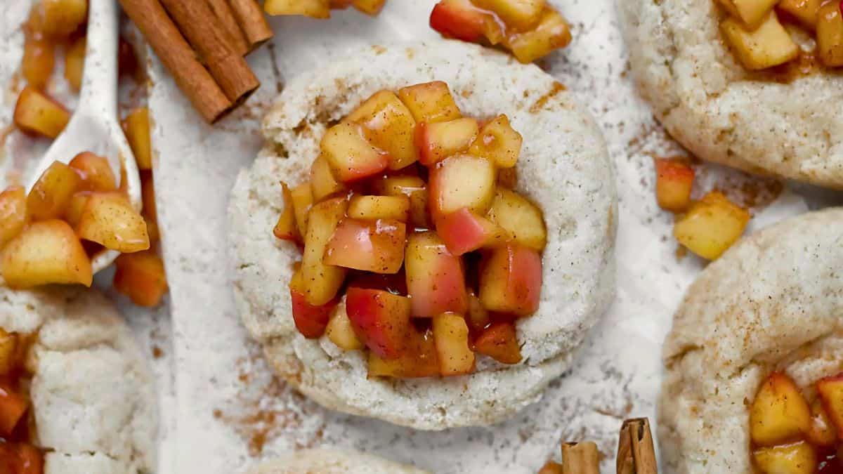 Close-up of a baked pastry topped with diced apples and cinnamon, surrounded by cinnamon sticks on a parchment paper. The apples are glazed, giving a shiny appearance, with spices sprinkled around the pastry.