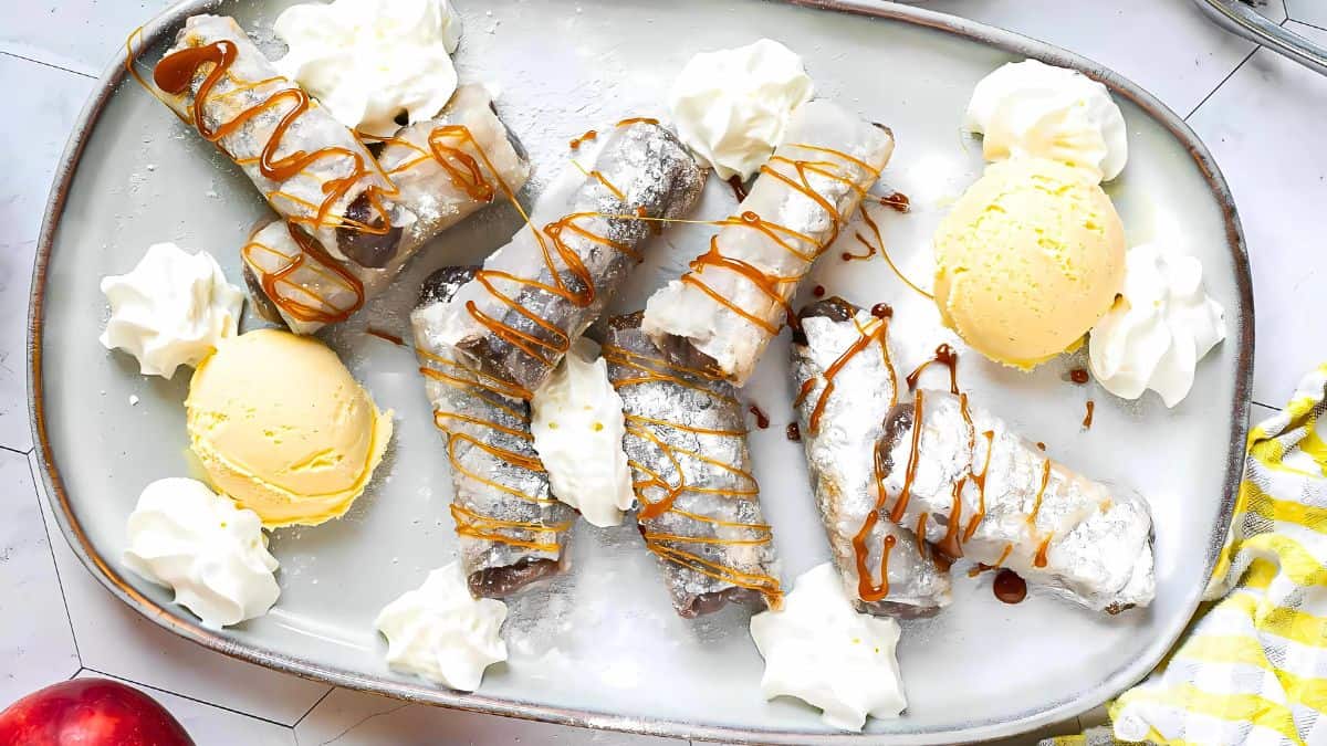 A plate of dessert spring rolls filled with likely chocolate, drizzled with caramel sauce, and dusted with powdered sugar. Accompanied by scoops of vanilla ice cream and dollops of whipped cream.