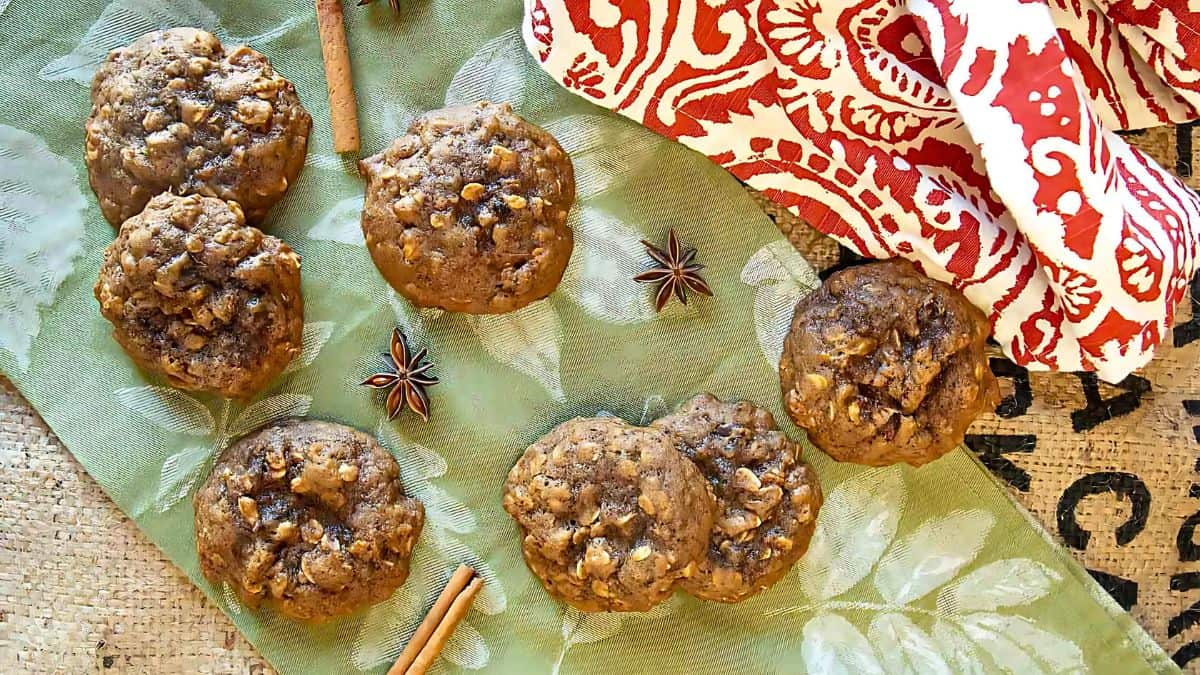 A set of seven cookies with nuts and chocolate chips is arranged on a green leaf-patterned surface. Nearby are star anise and cinnamon sticks, with a red and white patterned cloth draped above.