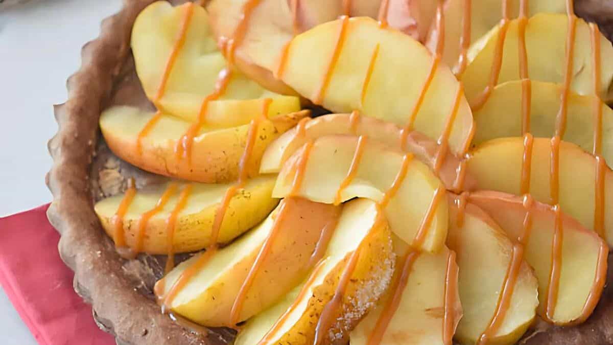 A close-up of a fruit tart with sliced apples arranged on a crust, drizzled with caramel sauce. The tart rests on a red napkin, showcasing a glossy caramel sheen over the apple slices.