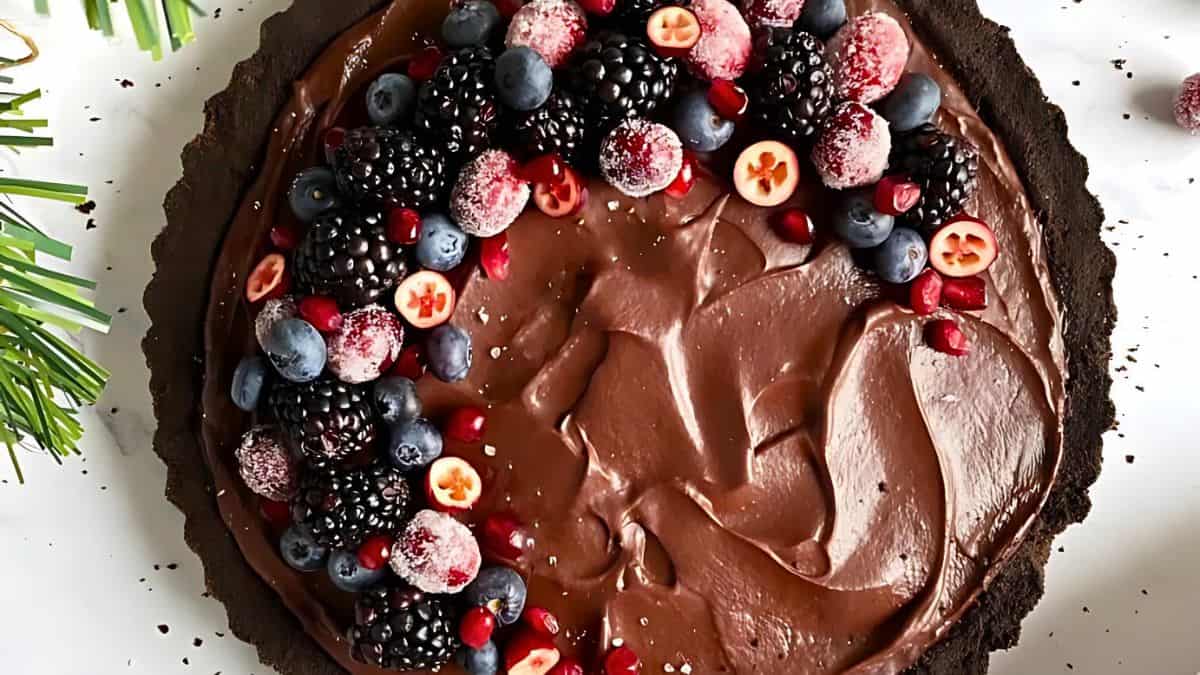 A chocolate tart topped with a variety of berries, including blackberries, blueberries, and frosted cranberries, arranged in a semi-circle. The tart rests on a white surface, decorated with small greenery on the side.