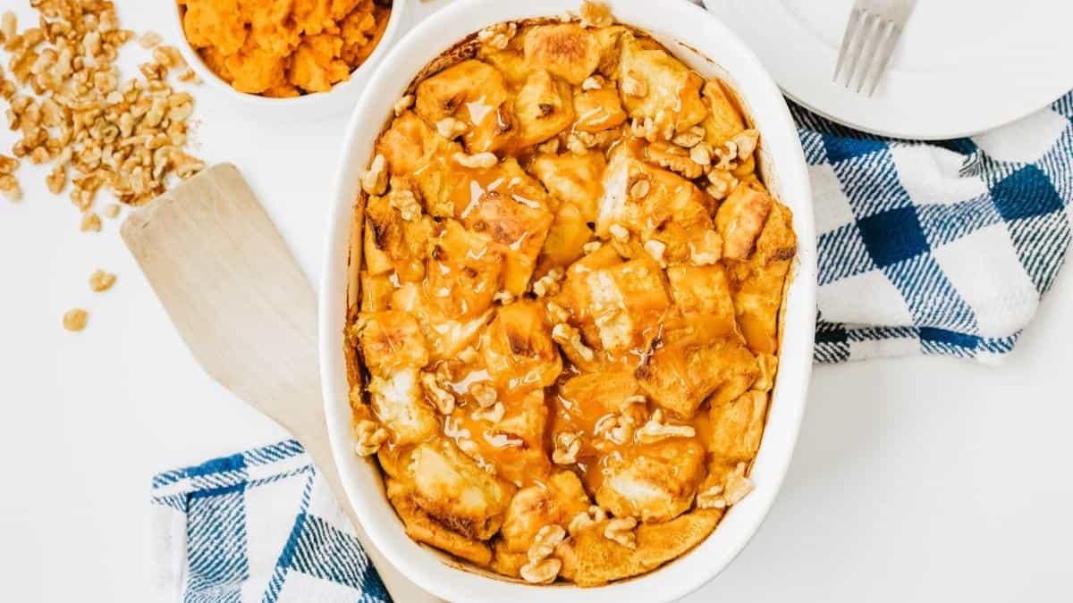 A baked dish filled with an orange bread pudding garnished with walnuts sits on a white table. A bowl of mashed sweet potatoes, a wooden spatula, and a blue checkered cloth are nearby.