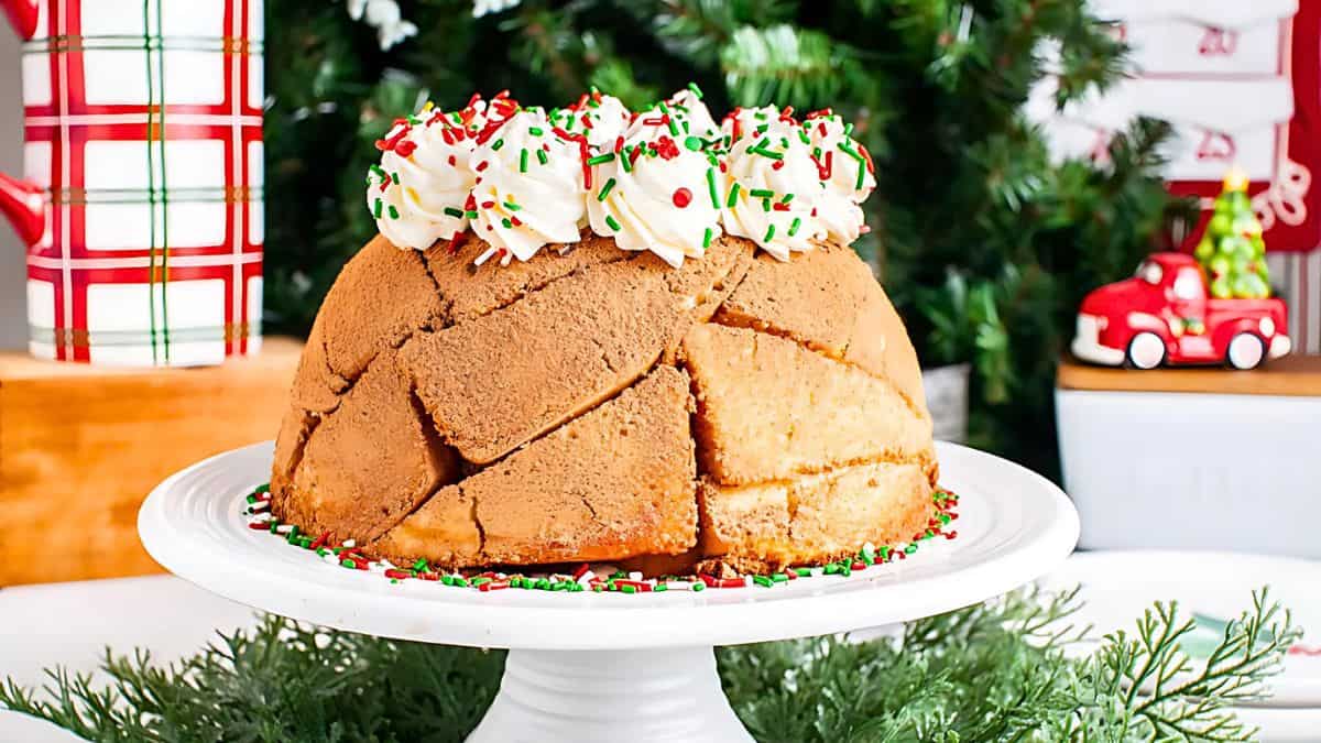 A festive dome-shaped cake on a white stand, topped with whipped cream and red and green sprinkles. The cake is surrounded by a sprinkle border. In the background, a decorated Christmas tree and holiday decor can be seen.
