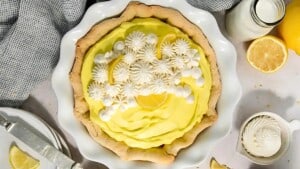 A lemon meringue pie with a golden crust is topped with piped whipped cream and lemon slices. It's set in a white pie dish, surrounded by a checkered cloth, lemon halves, and a glass jar of milk.