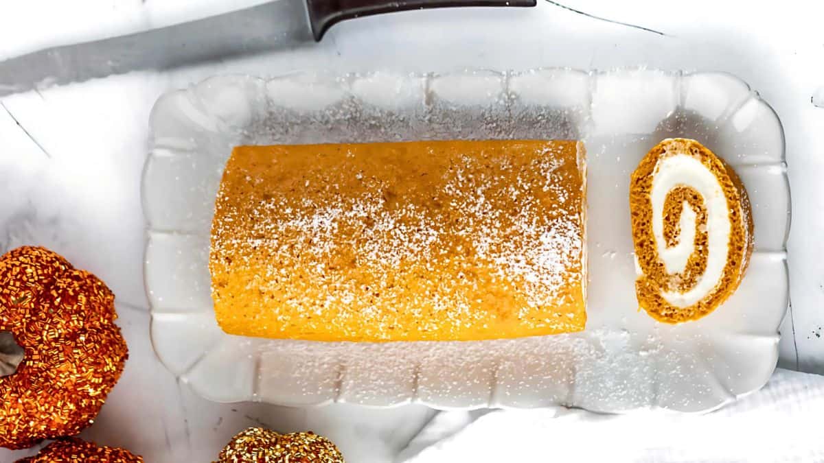 Pumpkin roll with cream filling on a rectangular glass platter, sprinkled with powdered sugar. A slice is cut and placed next to the roll. Nearby, there are decorative elements resembling mini pumpkins.