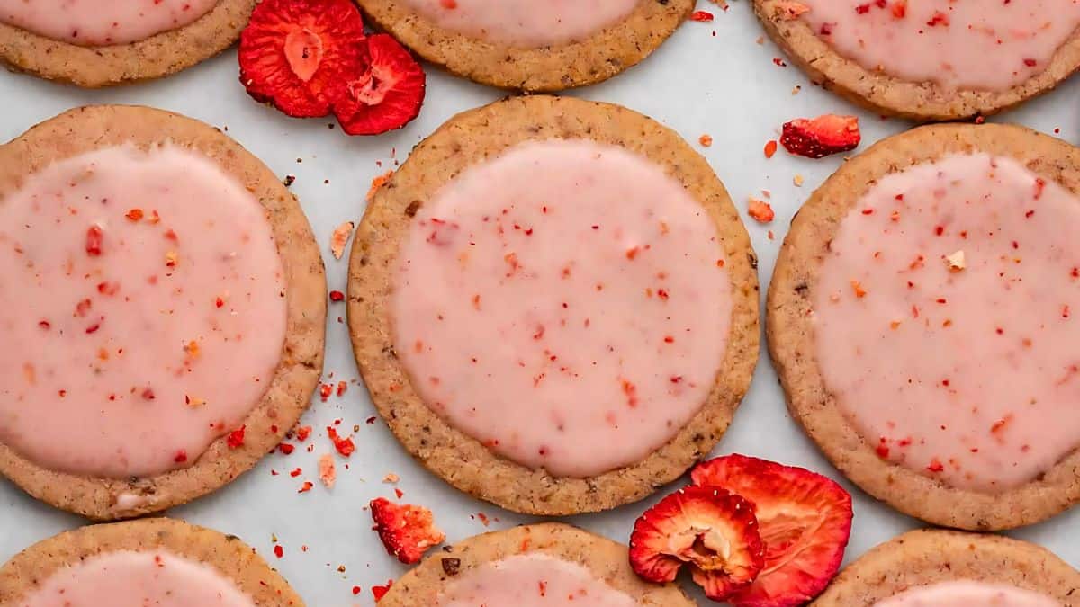 Cookies with a light pink icing are topped with small strawberry pieces. The round cookies are evenly spaced on a white background, and there are slices of fresh strawberries scattered among them.
