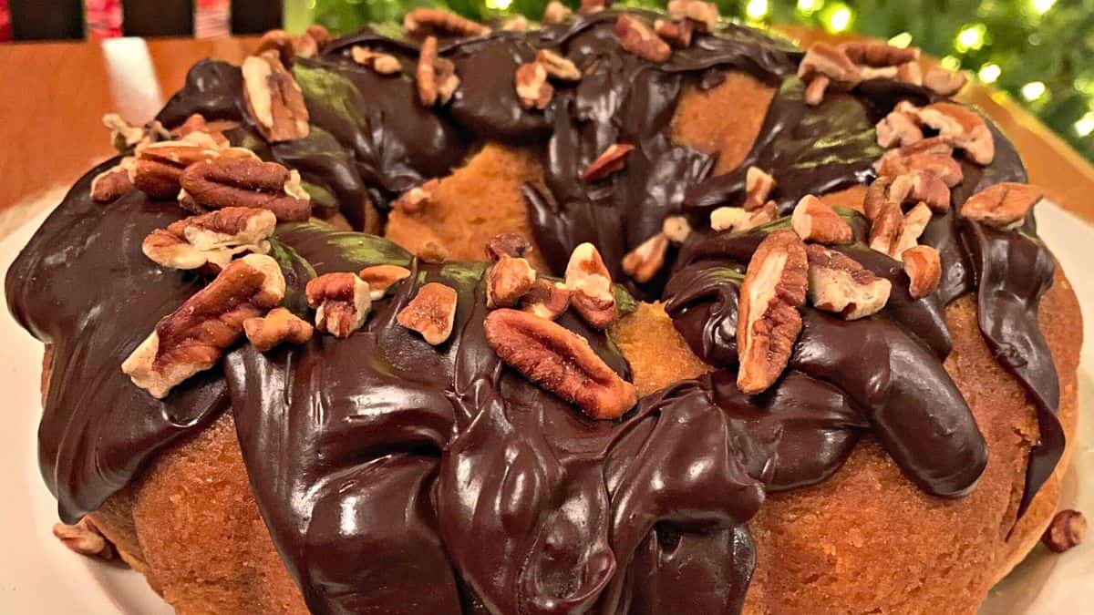 A bundt cake topped with rich, glossy chocolate icing and sprinkled with pecans, set against a warm, festive background.