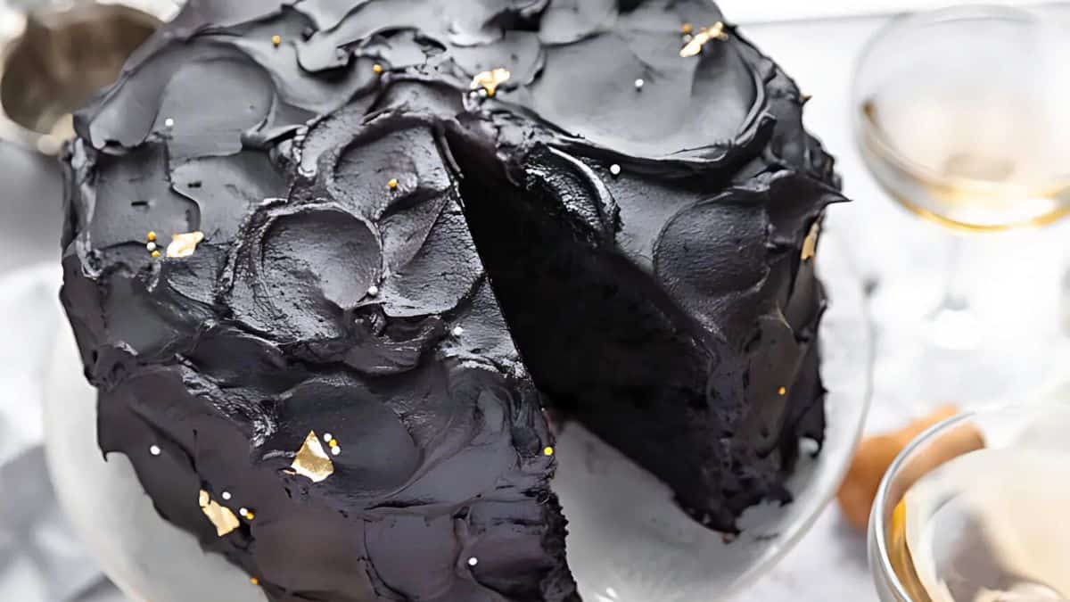 A round black cake with rich, glossy frosting, partially sliced to reveal its layers. Small gold flakes are sprinkled on top, adding a touch of elegance. The cake is on a glass plate near two champagne flutes.
