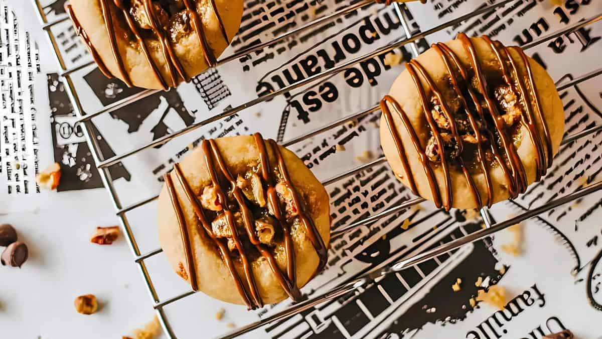 Cookies drizzled with chocolate and topped with nuts rest on a wire rack over newspaper wrappers. Chocolate chips are scattered nearby on the white surface.