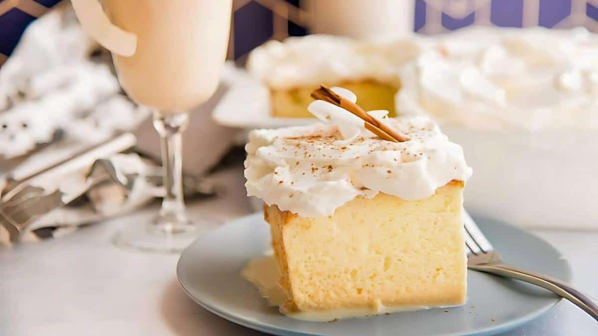 A slice of creamy dessert topped with whipped cream and a cinnamon stick on a blue plate. A glass of a frothy beverage and another serving of dessert with whipped topping are visible in the background.