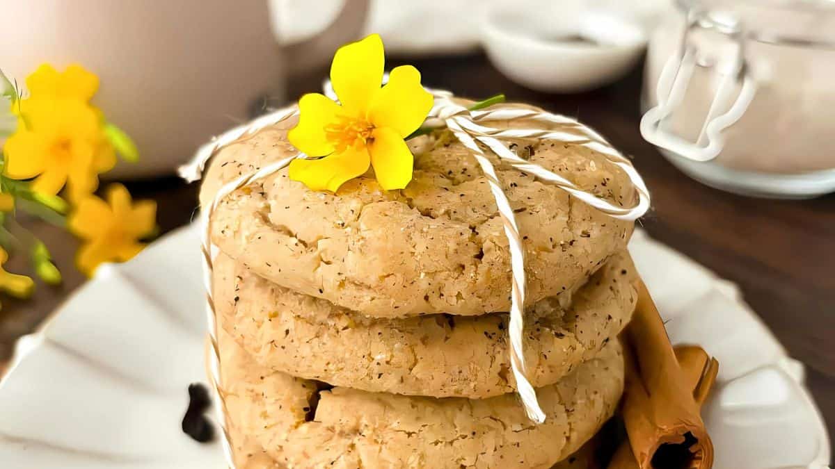 A stack of cookies tied with a string is adorned with a bright yellow flower. The cookies are on a white plate, with a cinnamon stick beside them. In the background, there’s a jar of sugar and additional yellow flowers for decoration.