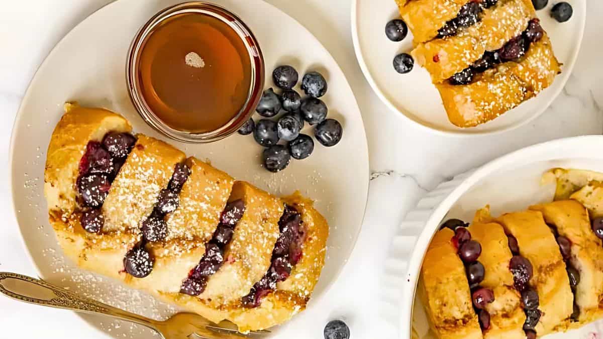 A plate featuring sliced French toast dusted with powdered sugar, topped with blueberries, and served with a small dish of syrup. Additional plates containing the same dish are nearby, along with extra blueberries scattered on the marble surface.