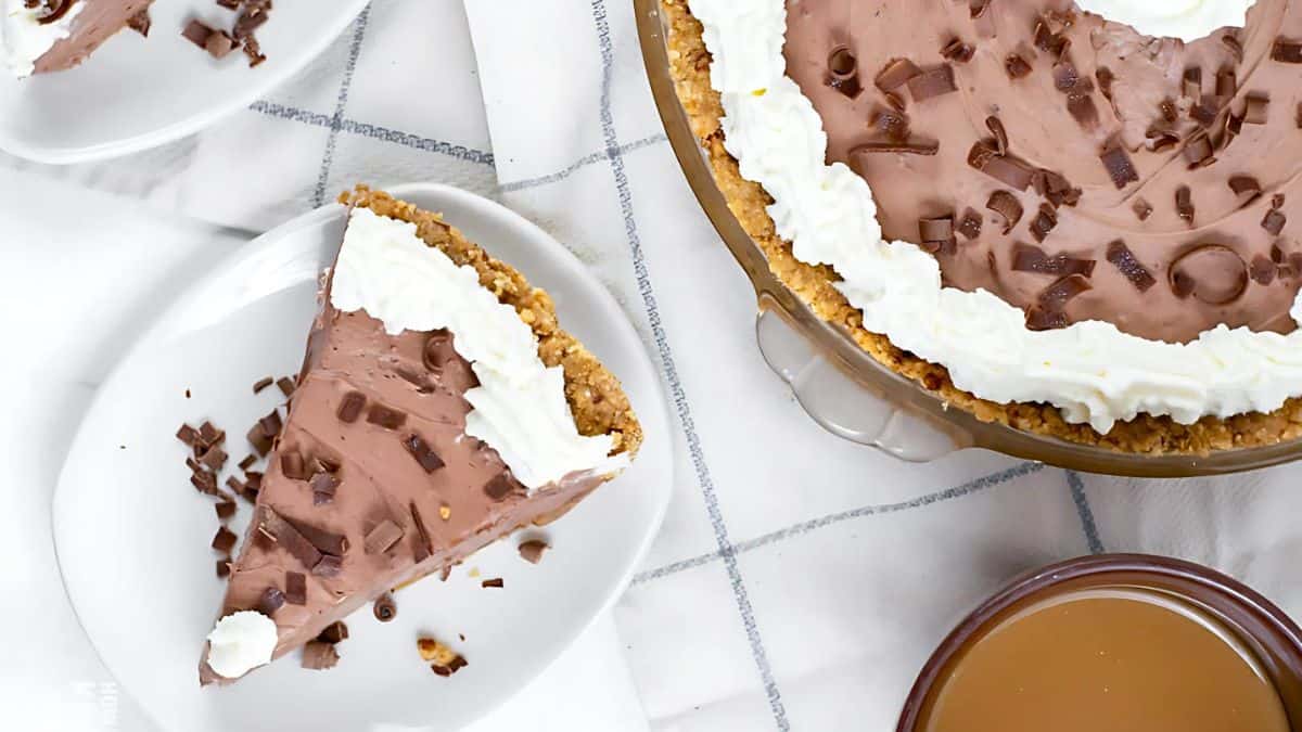 A chocolate cream pie with a graham cracker crust, topped with whipped cream and chocolate shavings. A slice is served on a plate beside the pie. There's a bowl of caramel sauce nearby on a white checkered tablecloth.