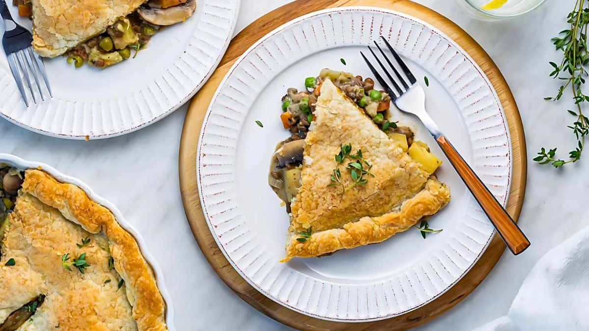 A table setting with two white plates, each holding a slice of savory pie filled with vegetables. The pies are topped with golden, flaky crust and garnished with fresh thyme, next to forks. A partly seen pie dish and a glass with lemon water are in the background.