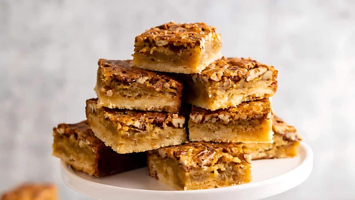 A stack of pecan pie bars on a white cake stand. The bars have a golden brown crust with a glossy, nut-filled topping, featuring a mix of crumbly and chewy textures. The background is softly blurred, highlighting the dessert.