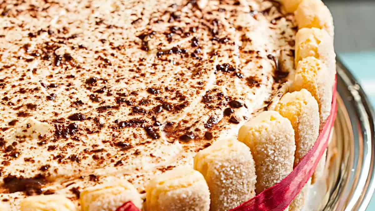 Close-up of a tiramisu cake decorated with ladyfingers around the edge, tied with a red ribbon. The top is sprinkled with cocoa powder, creating a marbled effect on the creamy surface. The cake sits on a glass serving plate.