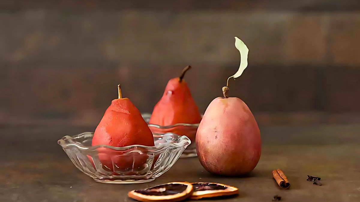 Three poached pears in small glass bowls sit on a dark surface. Two pears have stems, and one has a paper leaf attached. Slices of orange and a cinnamon stick add a touch of color in the foreground.