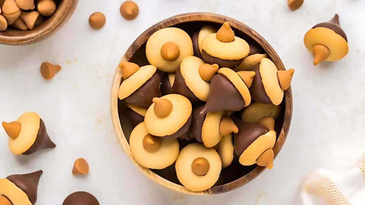 A wooden bowl filled with acorn-shaped treats made of cookies, chocolate kisses, and peanut butter chips. Some treats are scattered on the white surface around the bowl.