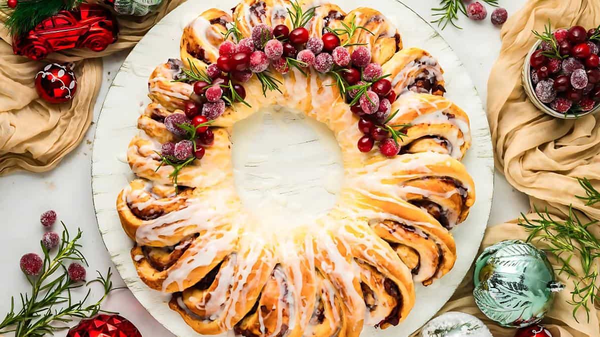 A festive bread wreath, adorned with white icing and garnished with sugared cranberries and rosemary sprigs. Surrounding decorations include red and green ornaments and beige fabric on a light wooden surface.