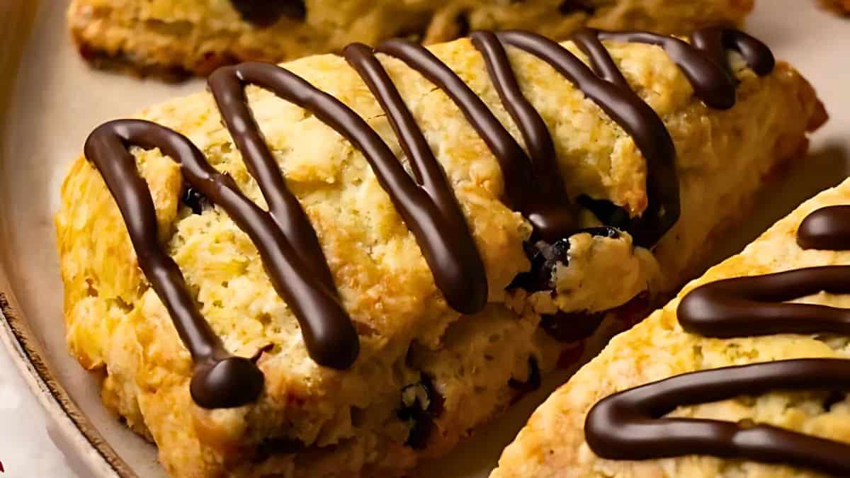 A close-up of a golden-brown scone drizzled with dark chocolate. The scone has a crumbly texture with small bits of dark filling, possibly chocolate or fruit, visible throughout. It's placed on a beige ceramic plate with other similar scones around it.