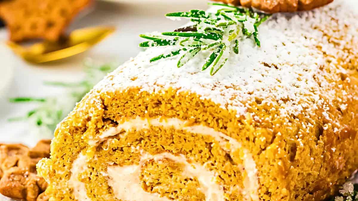 A pumpkin roll filled with cream cheese frosting, sprinkled with powdered sugar on top. It's garnished with a sprig of greenery and a small cookie. The roll is on a light-colored surface with a blurred background.
