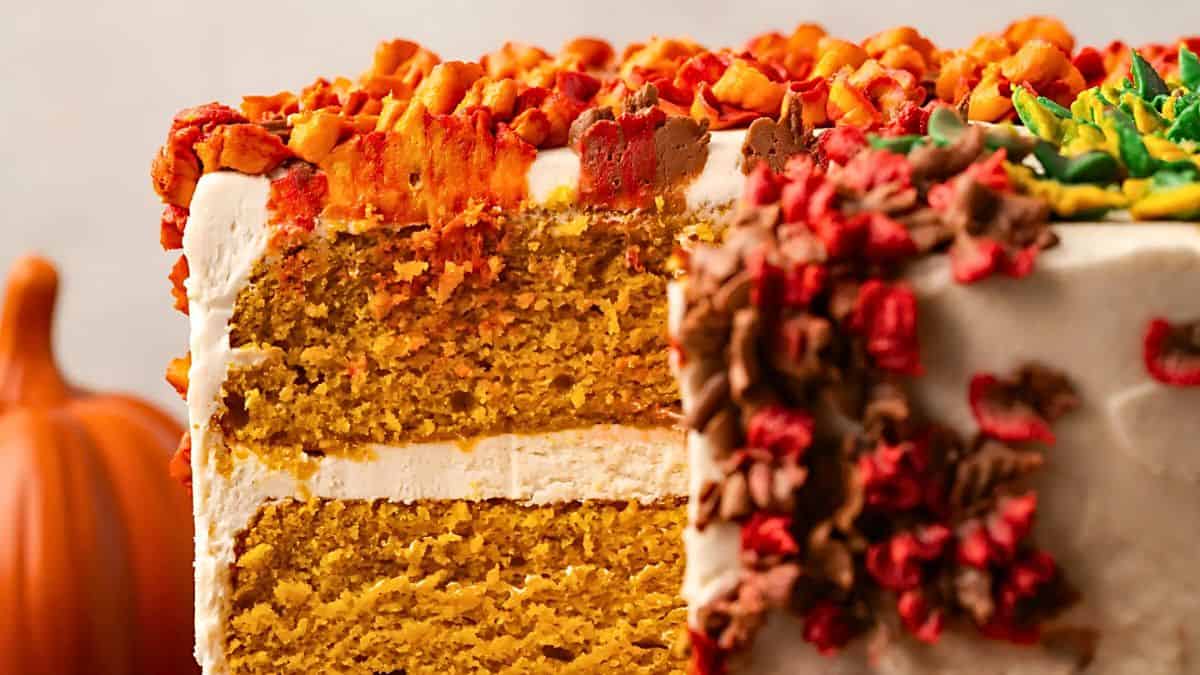 Close-up of a sliced pumpkin spice cake with creamy layers and colorful fall-themed frosting, topped with decorative toppings. A pumpkin is blurred in the background.
