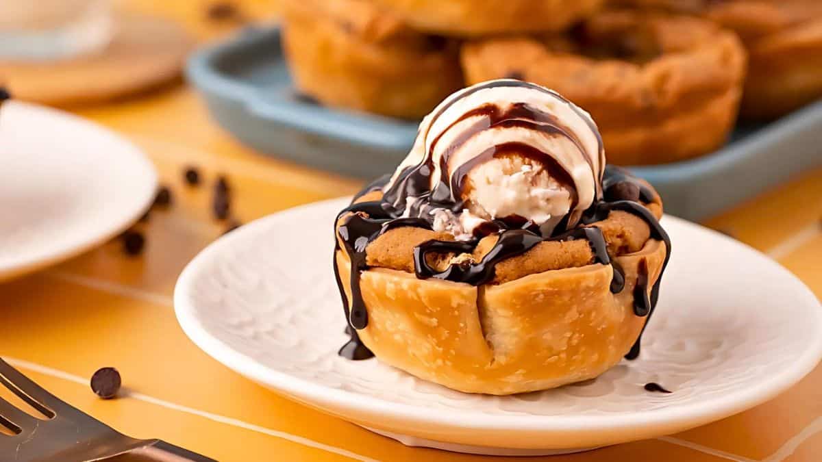 A mini pie on a white plate topped with a scoop of vanilla ice cream drizzled with chocolate sauce. In the background, more mini pies and chocolate chips are visible on a blue tray.