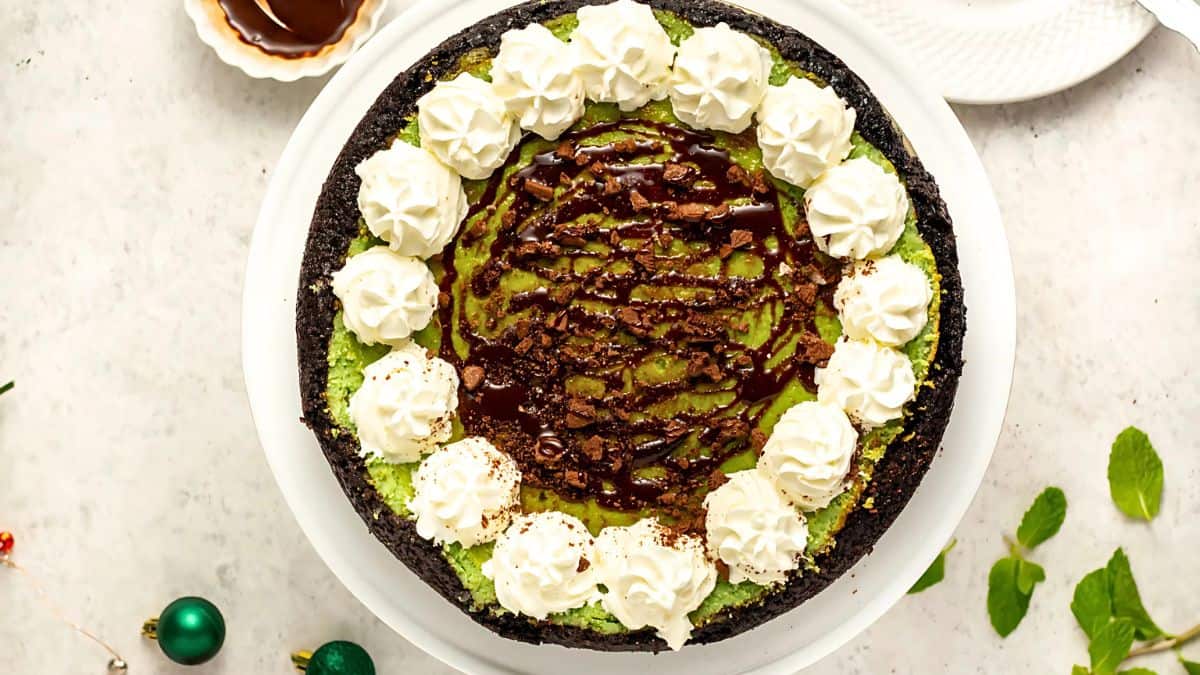 A top-down view of a green pie in a dark crust, topped with swirls of chocolate and a border of whipped cream dollops. The pie is on a round white plate. Some mint leaves and a small bowl of chocolate sauce are nearby.