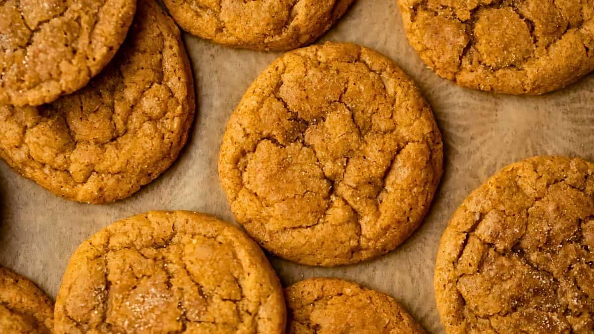 Close-up of several round, golden-brown cookies arranged on a parchment-lined surface. The cookies have a slightly cracked texture and appear freshly baked, suggesting a warm and inviting aroma.