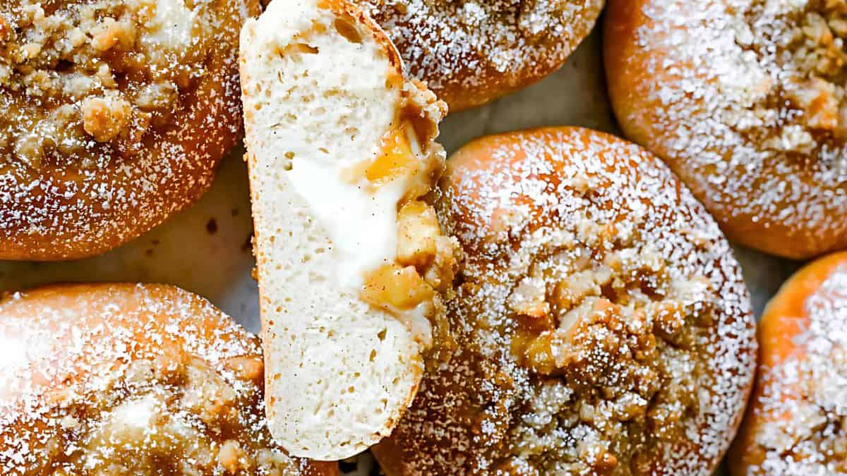 Close-up of apple-stuffed buns with crumb topping and powdered sugar. One bun is cut in half, revealing a creamy filling and apple chunks. The golden-brown buns are arranged closely on a tray.