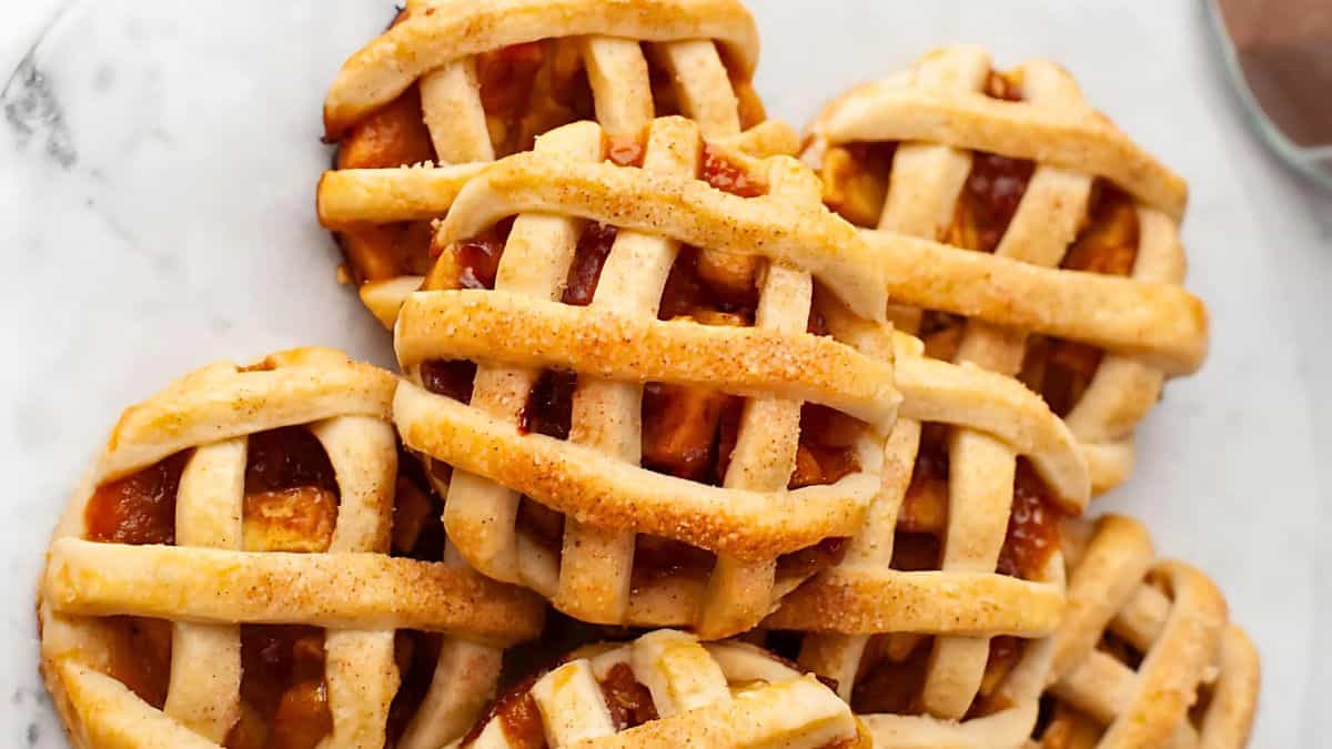 A plate of mini apple pies with lattice crusts, showcasing golden-brown tops and visible chunks of apple filling. The pies are arranged closely together on a white marble surface.