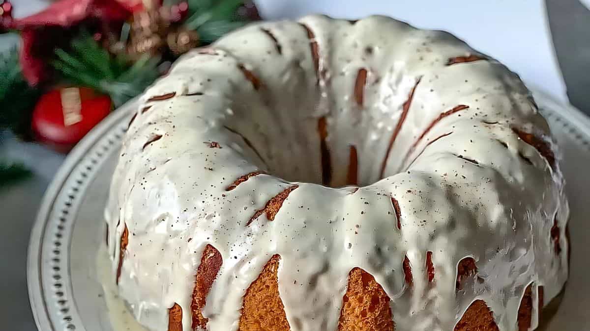A close-up of a Bundt cake drizzled with white icing on a white platter. The background has festive decorations, including red and green elements, suggesting a holiday theme.