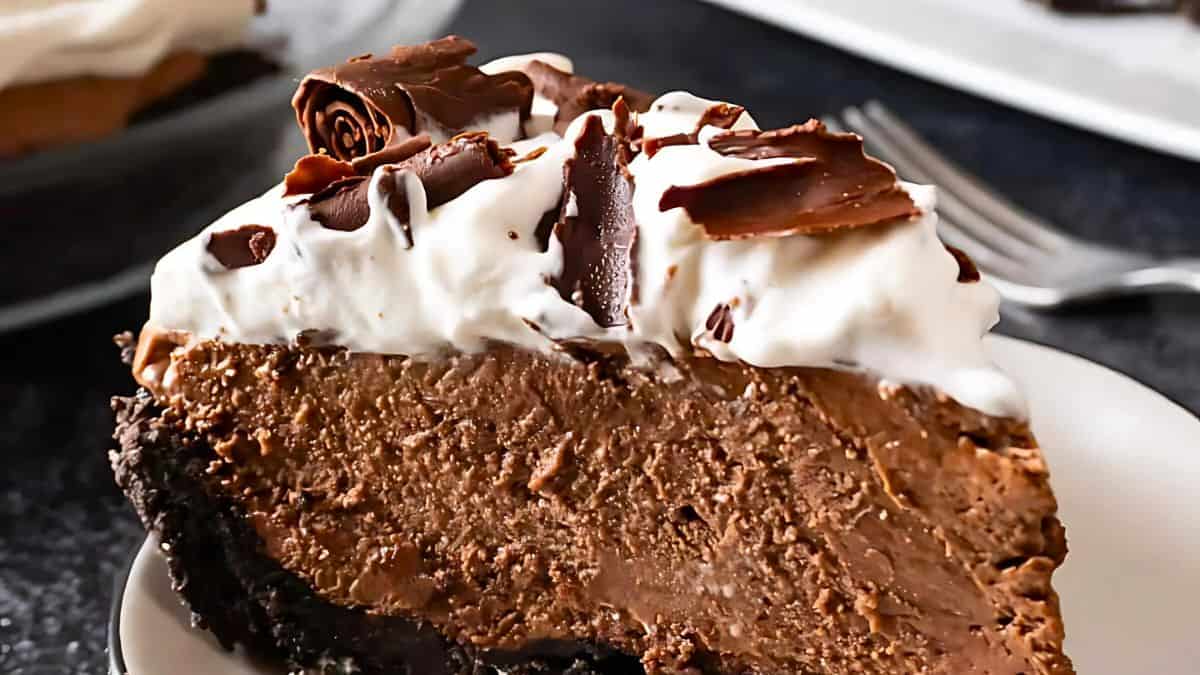 A slice of creamy chocolate pie with a dark crust, topped with whipped cream and chocolate shavings, served on a plate. A fork is placed nearby.
