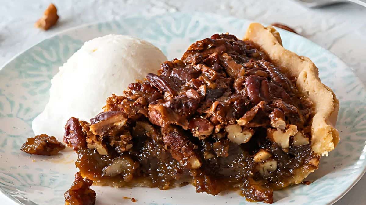 A slice of pecan pie on a plate, topped with caramelized pecans and served with a scoop of vanilla ice cream. The pie's crust is golden brown, and the filling is gooey and rich.