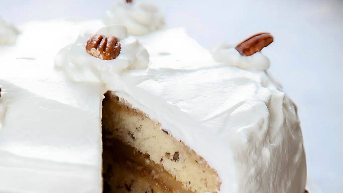 A close-up of a frosted cake with a slice cut out, showing layers of cream and filling. The cake is topped with smooth white icing and decorated with pecan halves.
