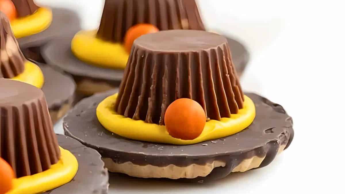 Close-up of a chocolate cookie treat with a peanut butter cup turned upside down on top, surrounded by yellow icing, and an orange candy resembling a pilgrim hat buckle. Several similar treats are visible in the background.