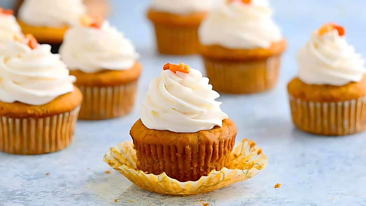 Pumpkin cupcakes with cream cheese frosting and small orange sprinkles are arranged on a light blue surface. One cupcake is in focus with its wrapper partially peeled. Other cupcakes are blurred in the background.