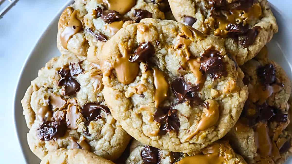 A plate filled with freshly baked cookies, each topped with melted chunks of chocolate and swirls of caramel. The cookies have a slightly golden-brown, chewy appearance.