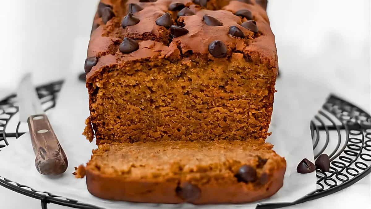 A loaf of pumpkin bread on a cooling rack, topped with chocolate chips. One slice is cut and lies in front, revealing a moist, dense texture. A knife with a wooden handle rests beside the loaf. The background is softly blurred.