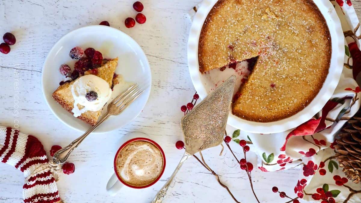 A golden-brown pie with a missing slice, garnished with cranberries, sits next to a slice topped with ice cream on a white plate. A fork rests on the plate. Nearby, a festive mug of coffee and a holiday cloth create a cozy setting.