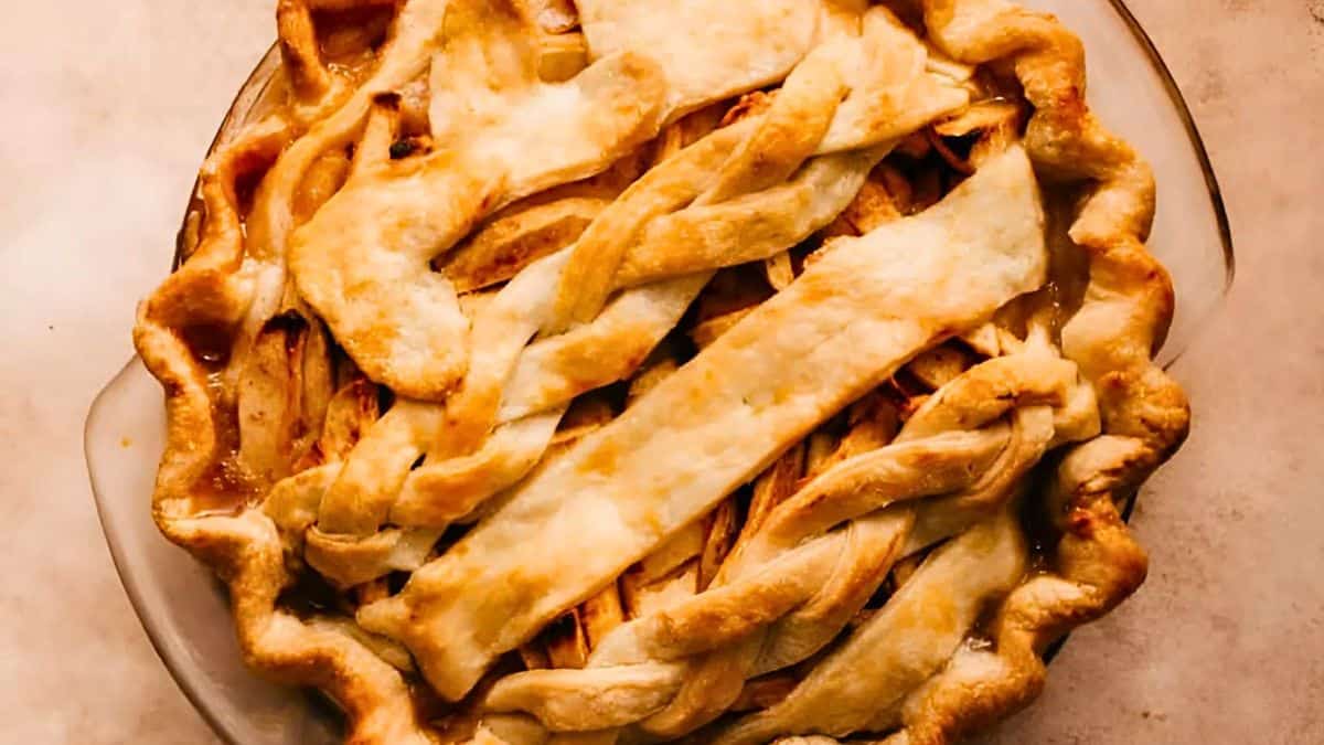 A freshly baked pie with a golden-brown, lattice crust, filled with visible slices of cooked apples, in a round, clear glass pie dish on a light countertop.