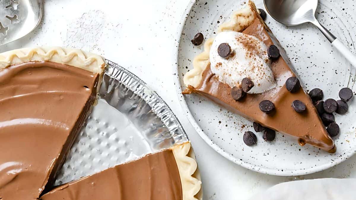 A creamy chocolate pie with a missing slice sits in a foil tray. The slice is served on a plate, topped with whipped cream, chocolate chips, and a dusting of cocoa. A spoon rests beside it on the speckled plate.